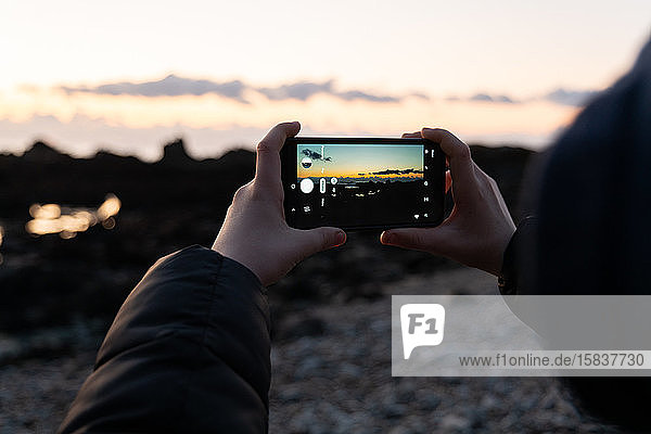Detail of hands holding smart phone taking photo of sunset over beach