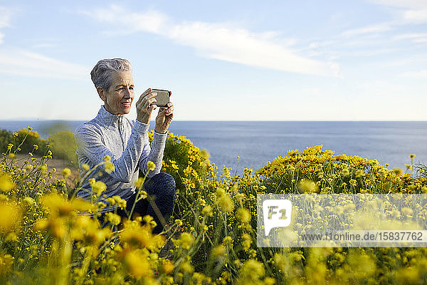 Ältere Frau fotografiert mit einem Smartphone  während sie sich bei Pflanzen gegen den Himmel hockt