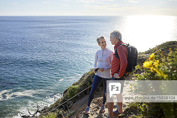 In voller Länge lächelndes älteres Ehepaar redet  während es an einem sonnigen Tag auf einer Klippe am Meer steht
