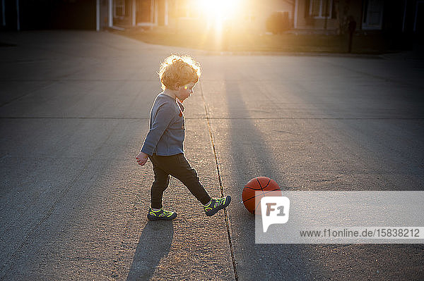Kleinkind Junge kickt Basketball draußen auf der Straße in ziemlich hellem Licht