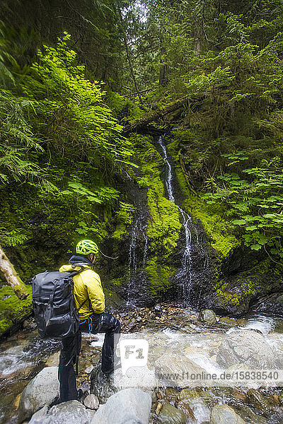 wet hiker wearing backpack looks at small waterfall.