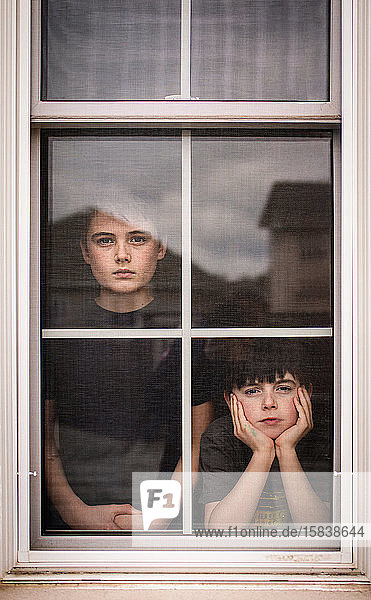 Two boys looking out of a window together with bored faces.