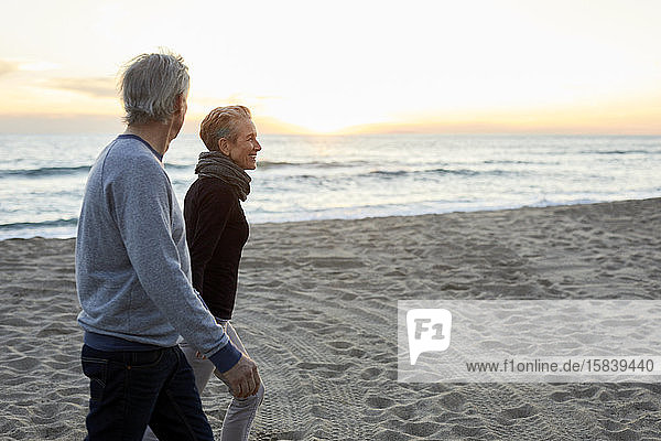 Seitenansicht eines lächelnden älteren Paares  das bei Sonnenuntergang am Strand spazieren geht