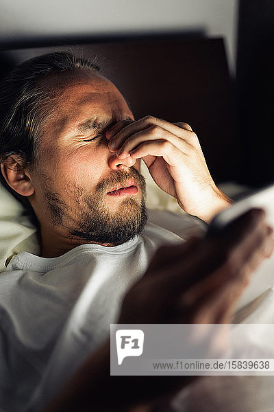 Bearded caucasian male with a headache in bed holding a tablet.