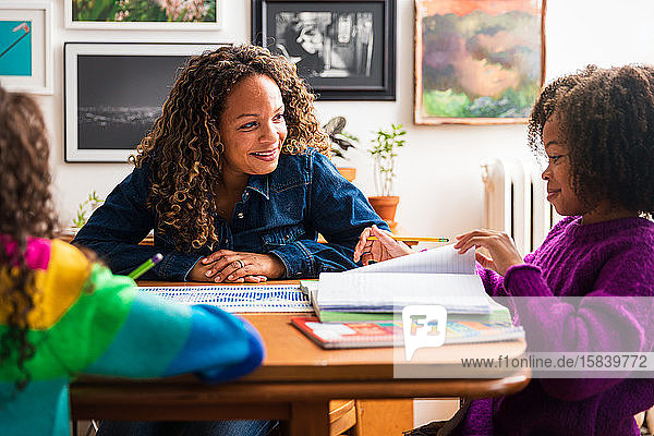 Lächelnde Mutter sieht Tochter beim Lernen am Tisch im Wohnzimmer an