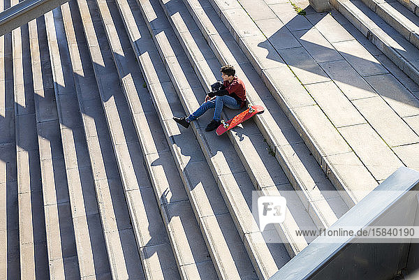 Junger Teenager sitzt auf einer Treppe im Freien und schaut bei Sonnenschein in die Kamera