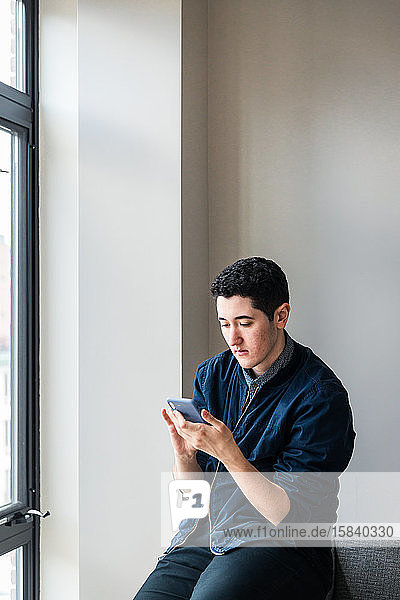 Junger Geschäftsmann benutzt Smartphone  während er im Büro auf dem Sofa an der Wand sitzt