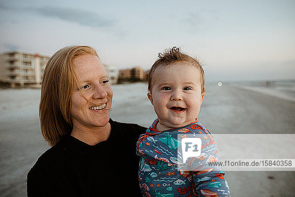 fat baby boy with crooked smile held by young red haired mom on beach