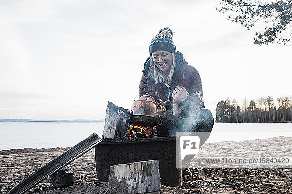 glückliche Frau  die sich am Strand am Lagerfeuer einen Wasserkocher aufwärmt