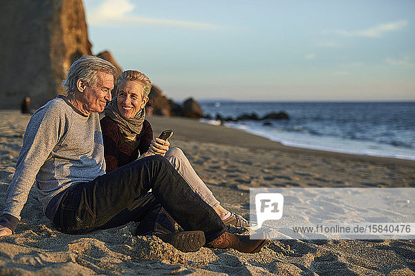 Älteres Ehepaar telefoniert  während es bei Sonnenuntergang am Strand im Sand sitzt