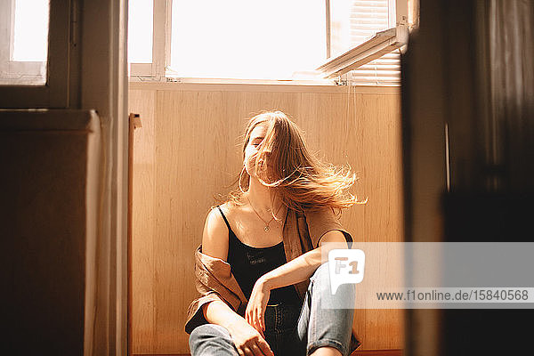 Young woman tossing hair while sitting on floor in balcony at home