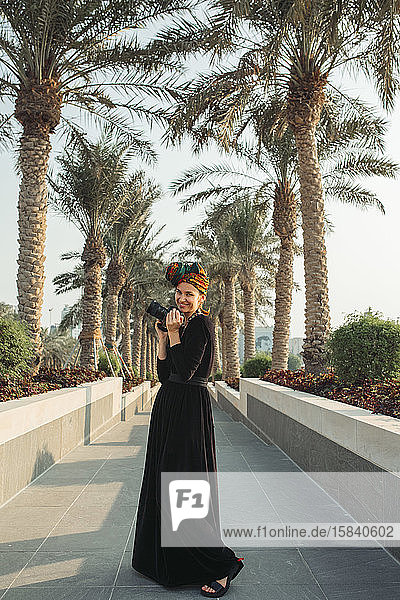 Junge Frau steht in einer Palmenallee und hält ihre Kamera
