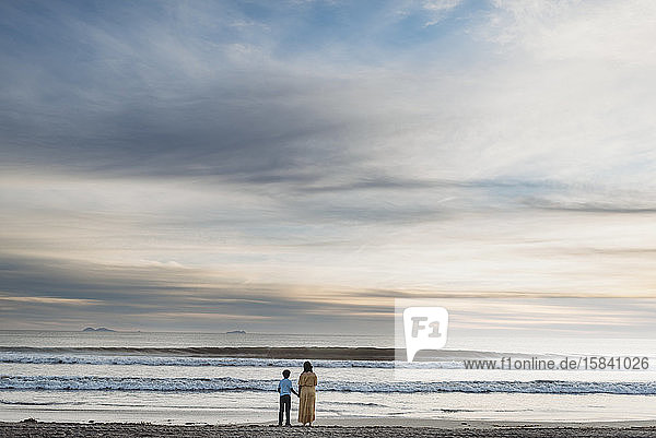 Junge Mutter mit Kleid und Sohn stehen bei bewölktem Sonnenuntergang am Strand