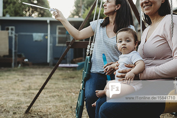 Two moms with baby on backyard swing playing with bubbles