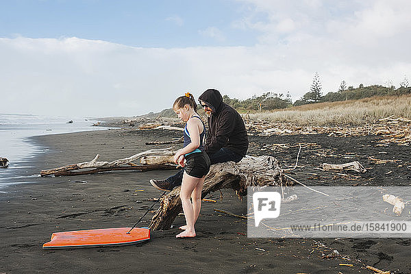 Man sitting on driftwood with girl and her boogie board on the beach