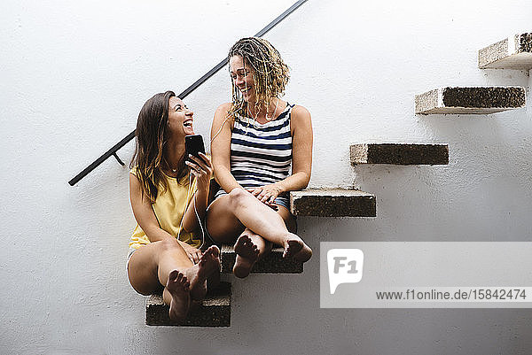 Zwei Frauen sitzen auf einer Treppe und lachen  während sie Musik hören.
