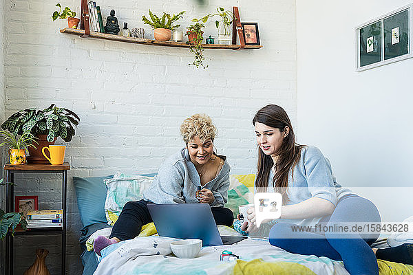 Frau zeigt Laptop ihrer Mitbewohnerin beim Kaffee trinken,  während sie zu Hause auf dem Bett sitzt