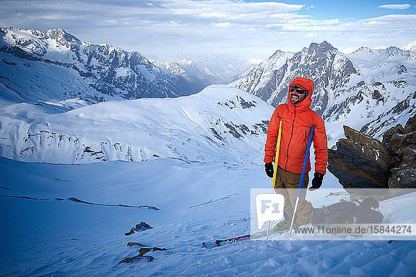 Skifahrer in orangefarbener Jacke  der sich auf Stöcke stützt und lacht
