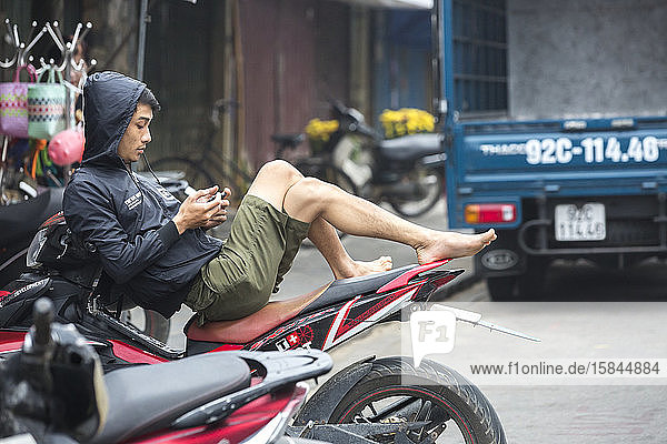 Vietnamesischer Mann entspannt sich auf Motorrad und schaut auf Smartphone.