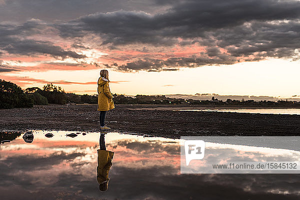 Girl in yellow coat standing on beach at sunset with reflection