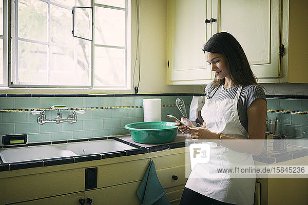 Junge Frau benutzt Mobiltelefon  während sie an der Küchentheke am Fenster steht