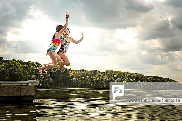 Zwei junge Mädchen in Badeanzügen springen von einem Dock in einen See