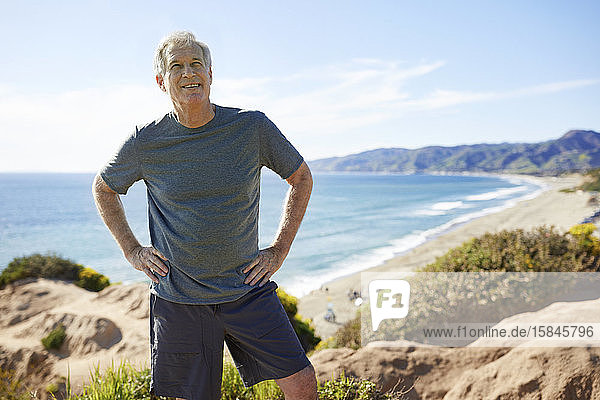 Smiling senior man with hands on hips standing on cliff at beach against sky