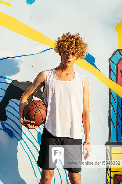 Young men with curvy hairs holding basketball ball in front of graffiti