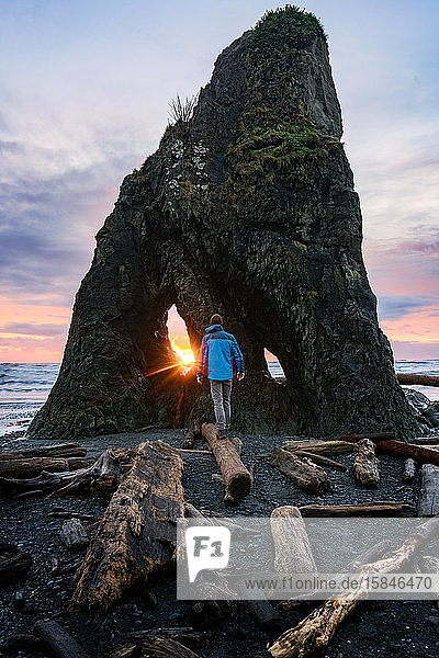 Mann geht auf einem Baumstamm am Strand bei Sonnenuntergang in der Nähe eines dramatischen Felsens