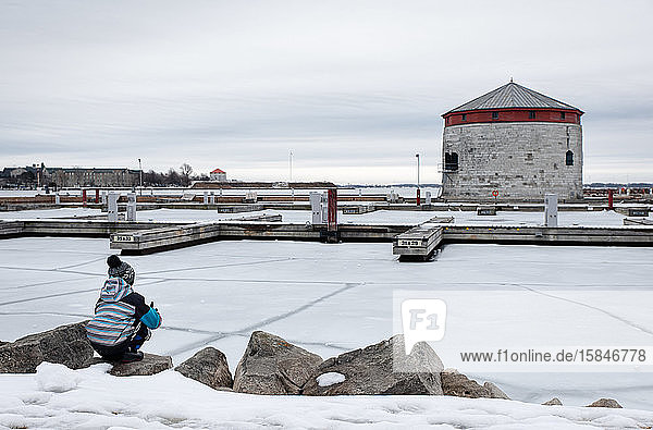 Boy sitting on edge of frozen lake looking at docks in the distance.