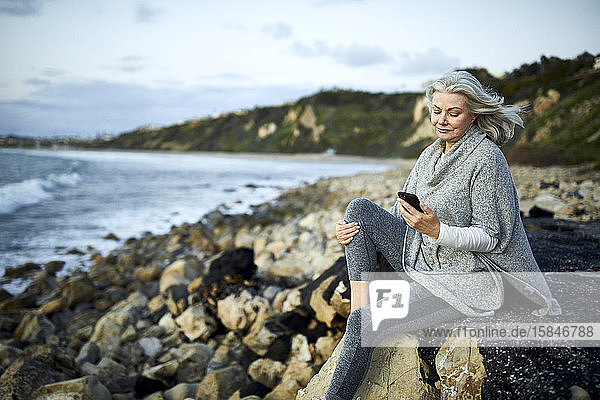 Ältere Frau benutzt ein Smartphone  während sie bei Sonnenuntergang auf einem Felsen am Strand von Manhattan gegen den Himmel sitzt