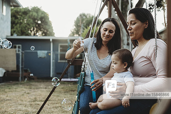 Baby girl on swing with two moms watching bubbles