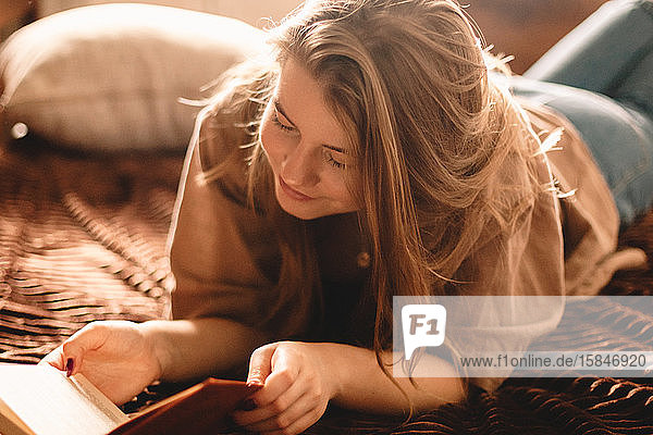 Junge glückliche Frau liest zu Hause auf dem Bett liegend ein Buch