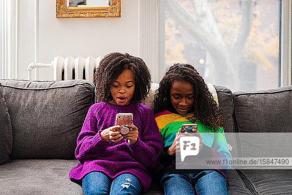 Schwestern in Pullovern  die zu Hause auf dem Sofa sitzen und dabei Mobiltelefone benutzen