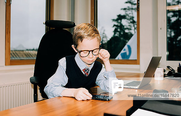 Junge  der in einem Büro arbeitet und bei der Arbeit auf das Telefon seines Vaters schaut
