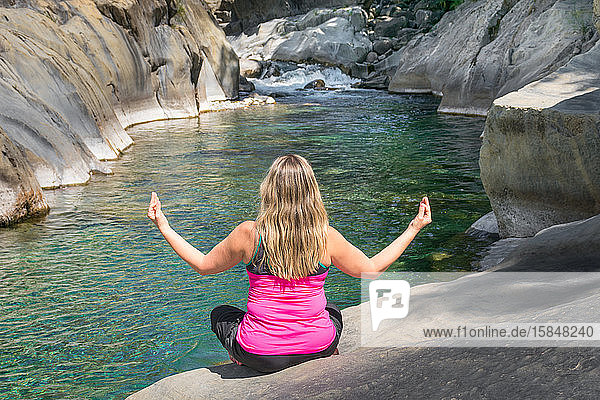 Frau rückwärts in sportlicher Kleidung  die an einem idyllischen Ort neben einem Fluss und einem Wasserfall zwischen weißen Felsen entspannende Yoga-Übungen praktiziert. Der Yogi sitzt mit erhobenen Armen auf dem Boden