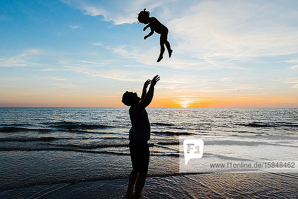 father throws his daughter into the air at sunset on beach vacation