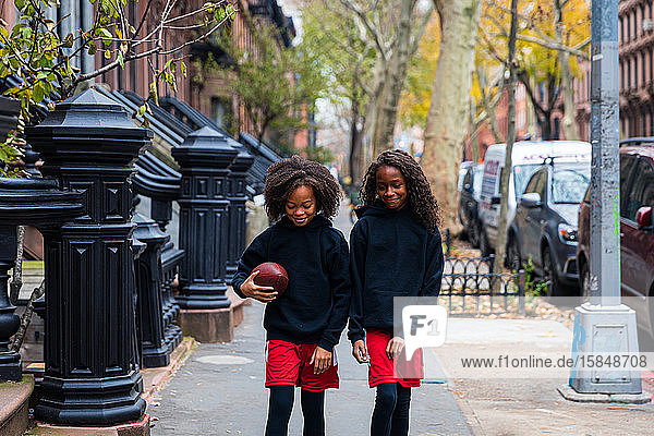 Mädchen hält American Football in der Hand  während sie mit ihrer Schwester auf einem Fußweg durch die Stadt läuft