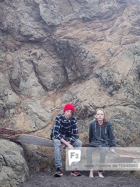 Teen brothers sitting on driftwood at the base of rocky cliff