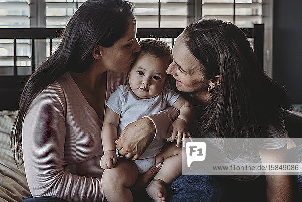 Two happy moms cuddling and kissing baby girl on bed near window