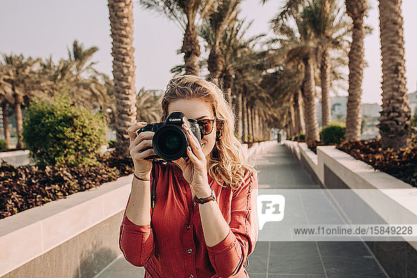 Junge Frau  die in einer Palmenallee steht und mit ihrer Kamera fotografiert