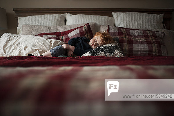 Toddler boy asleep on red plaid bed in pillows at home