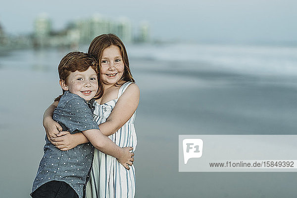 Porträt von Bruder und Schwester  die sich in der Abenddämmerung am Strand umarmen