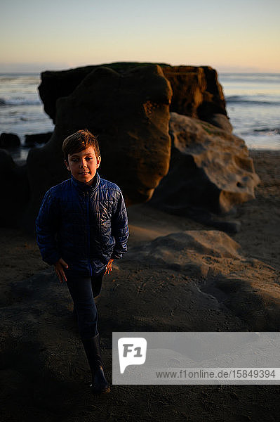 Boy standing near a boulder on the beach at sunset
