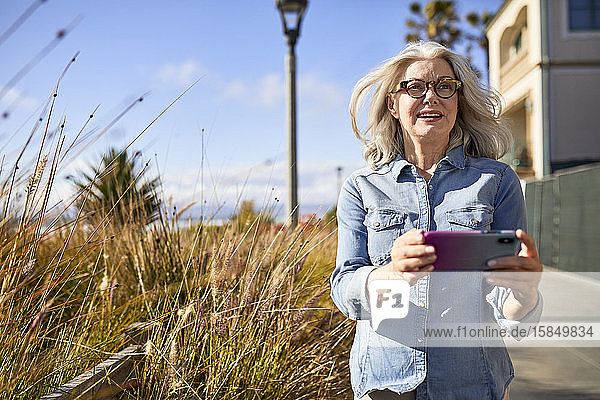 Lächelnde ältere Frau hält Handy in der Hand und schaut weg  während sie am Manhattan Beach steht
