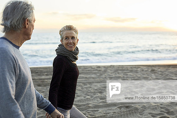 Seitenansicht eines älteren Paares  das sich beim Spaziergang am Strand bei Sonnenuntergang an den Händen hält