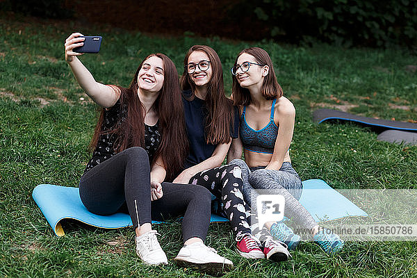 Drei Mädchen sitzen auf einer Yogamatte und machen Selfies im grünen Park