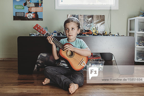 Pensive young boy sitting on hardwood floor with guitar