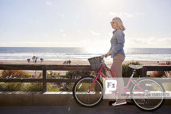 Seitenansicht einer älteren Frau mit Fahrrad  die am Manhattan Beach am sonnigen Tag auf der Straße gegen den Himmel steht