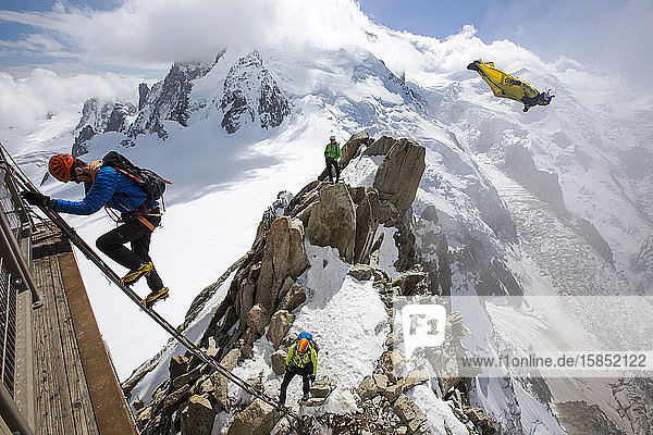Mont-Blanc von der Aiguille Du Midi oberhalb von Chamonix  Frankreich  mit Bergsteigern auf der Cosmiques Arete  die die Leiter zur Seilbahnstation erklimmen  und einem Base Jumper in einer Wing-Suite beim Fliegen